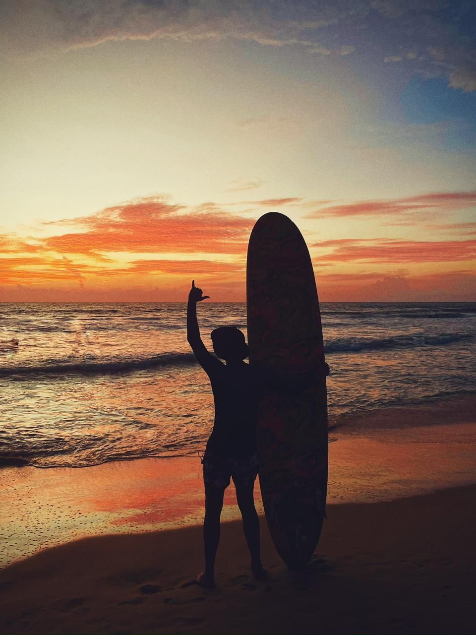 A surfer at dusk with a longboard standing upright in the sand, one arm raised