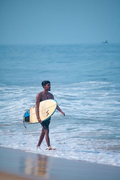 Movin walking the shoreline at Panambur with a yellow and blue board under his arm, the leash trailing behind him