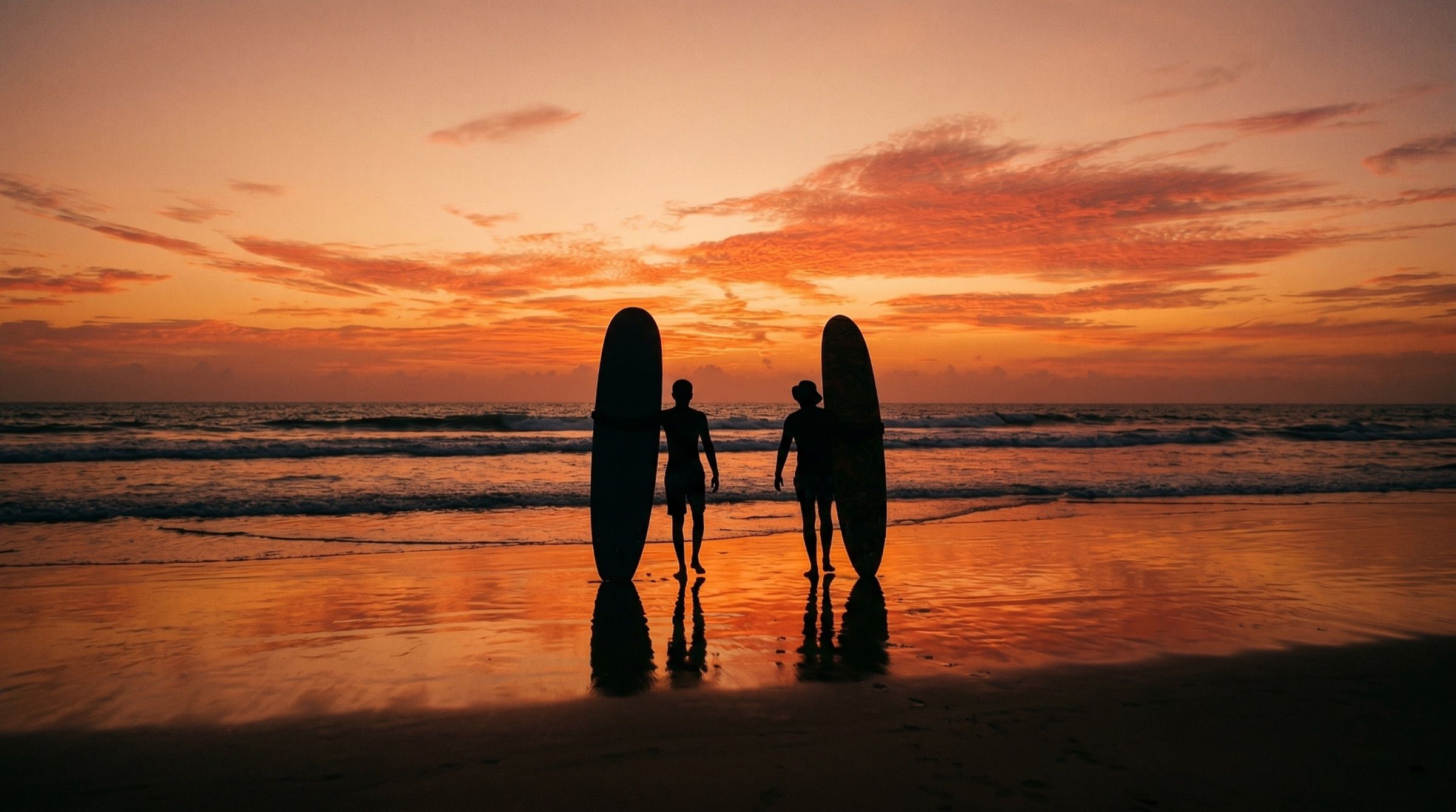 Two surfers standing in the shallows holding longboards, silhouetted against an orange sunset