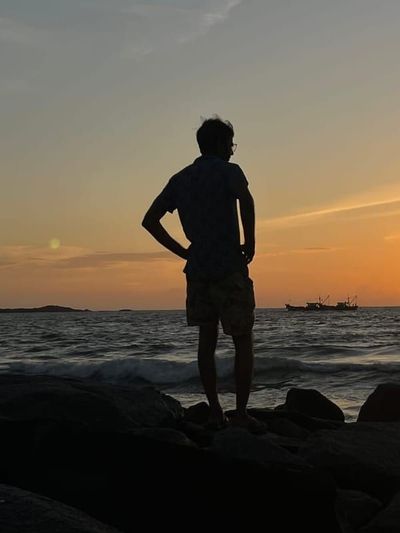 One person silhouetted on dark rocks at sunset, hands on hips, a fishing boat on the horizon and the sun low over the water