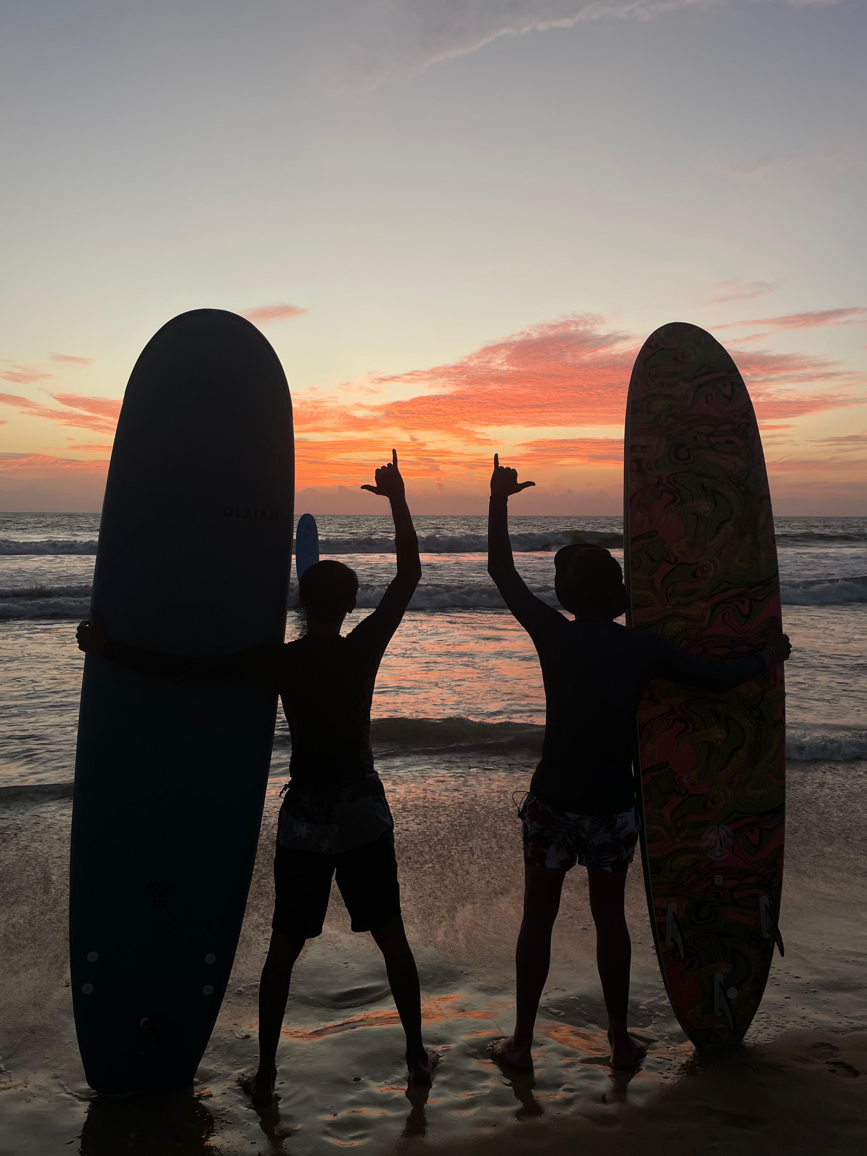 Two surfers on the sand at sunset with their arms up and longboards standing beside them