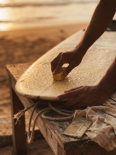 A hand rubbing a bar of wax on a longboard resting on a wooden bench at sunset, a coiled rope and a wax comb beside it